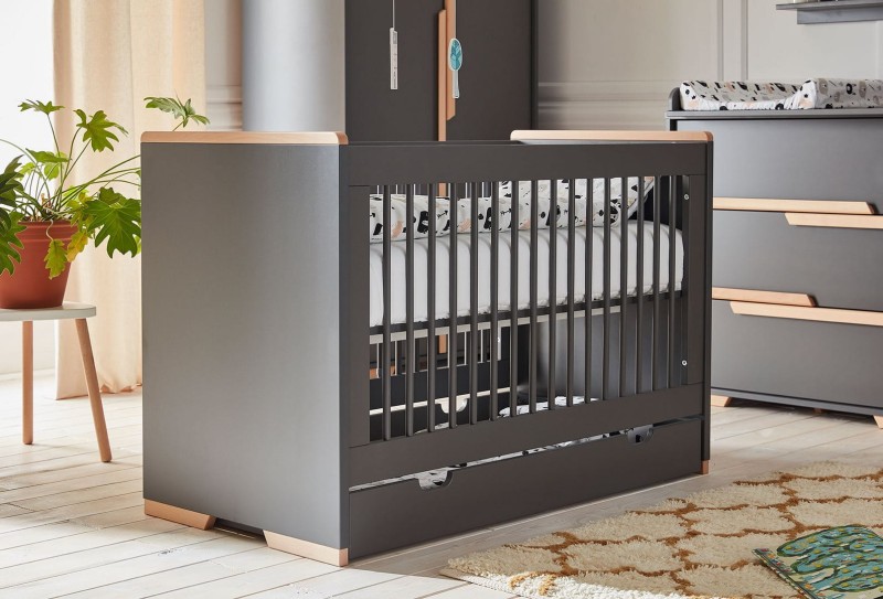 Szare łóżeczko dziecięce z szufladą i drewnianymi elementami w nowoczesnym pokoju.