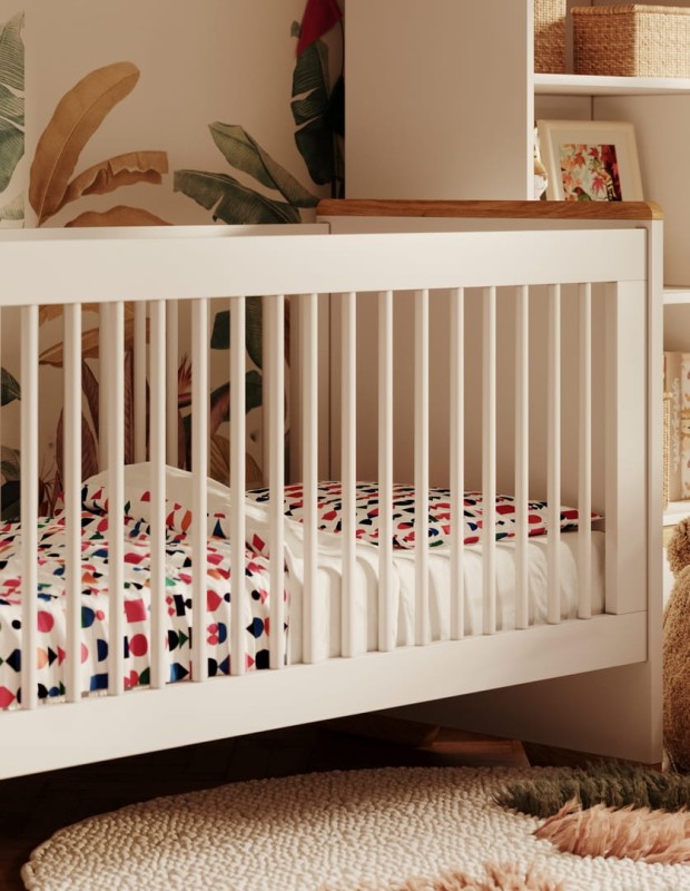 Zbliżenie na białe łóżeczko niemowlęce z kolorową pościelą na tle tapety w liście.