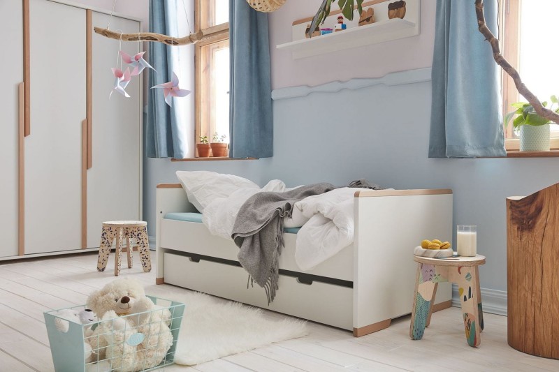 Białe łóżko dziecięce z szufladami i drewnianymi detalami, w aranżacji pokoju z zabawkami.