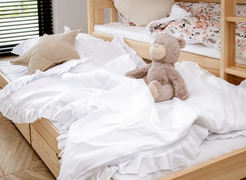 Przytulne łóżko dziecięce z białą pościelą z falbankami i pluszowym misiem.