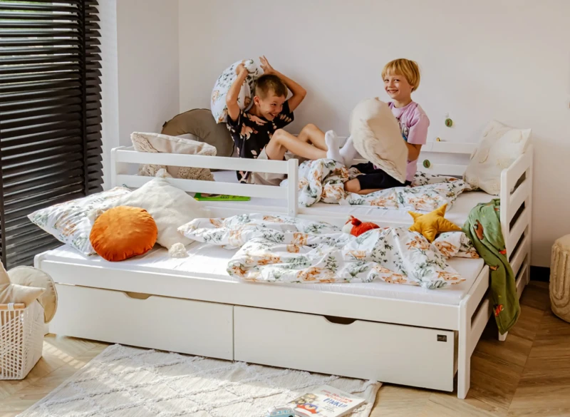 Dwoje radosnych chłopców podczas bitwy na poduszki na białym łóżku wysuwanym.