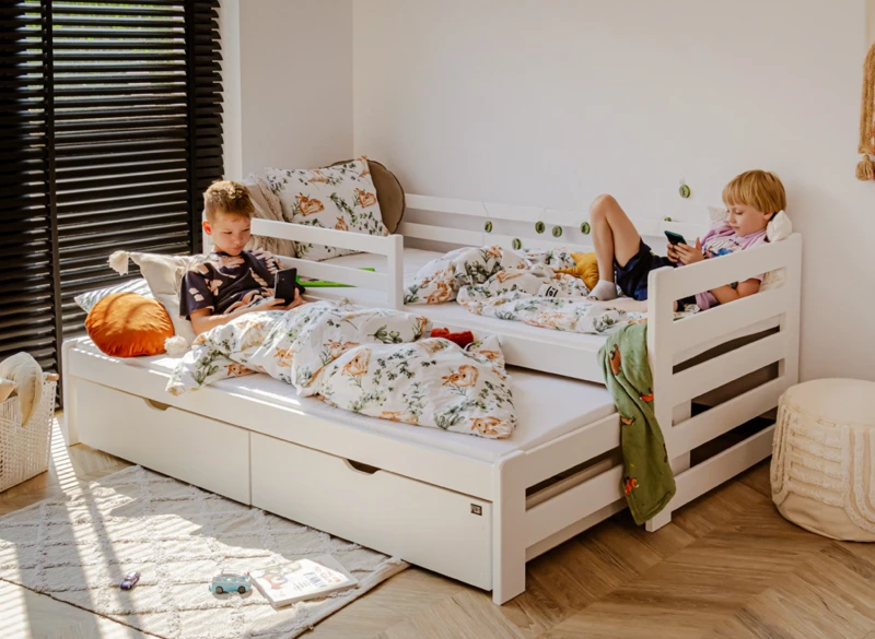 Dwóch chłopców na białym łóżku piętrowym z wysuwanym spaniem i pościelą w sarenki.