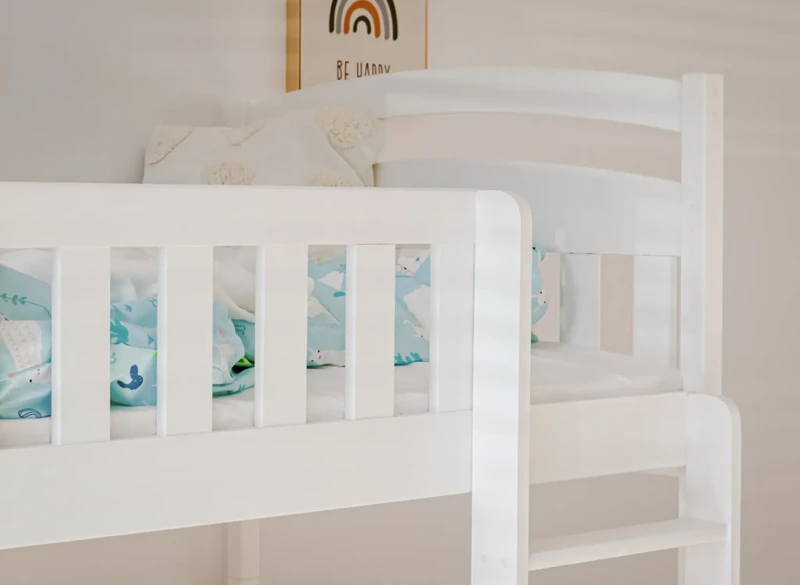 Zbliżenie na białą drewnianą barierkę górnego łóżka piętrowego z kolorową pościelą w tle.