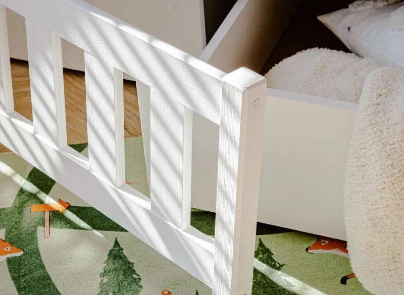 Detal białej barierki ochronnej łóżka na tle zielonego dywanu w lisy.