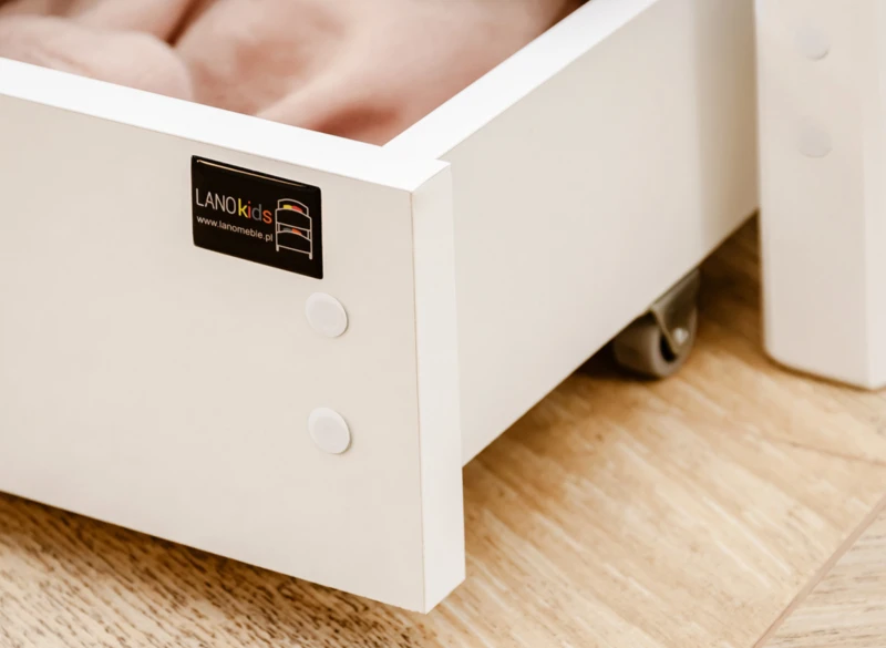 Detal białej szuflady pod łóżkiem z logo Lano Kids na kółkach.