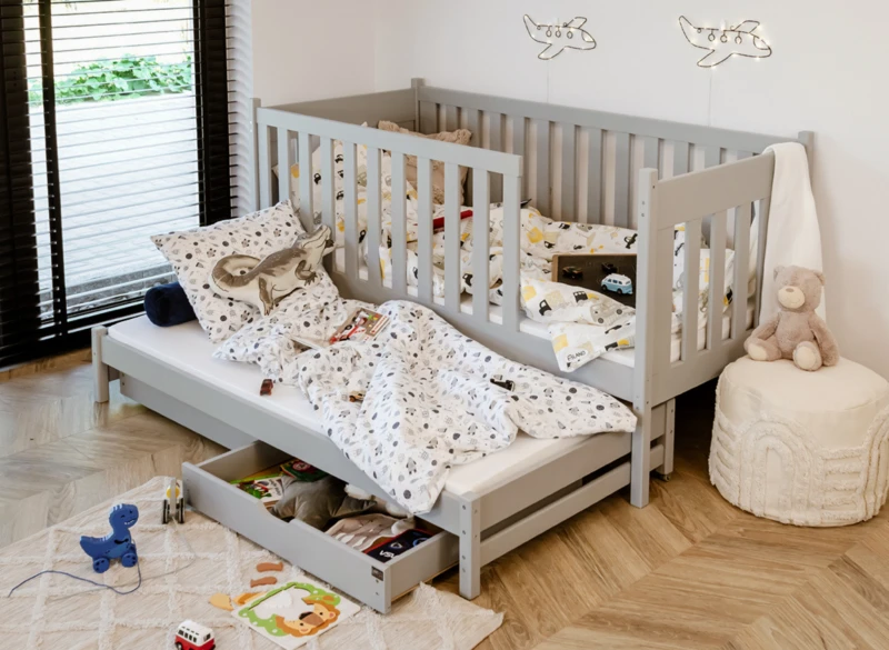 Szare łóżko dziecięce z barierką i wysuwanym dolnym materacem.