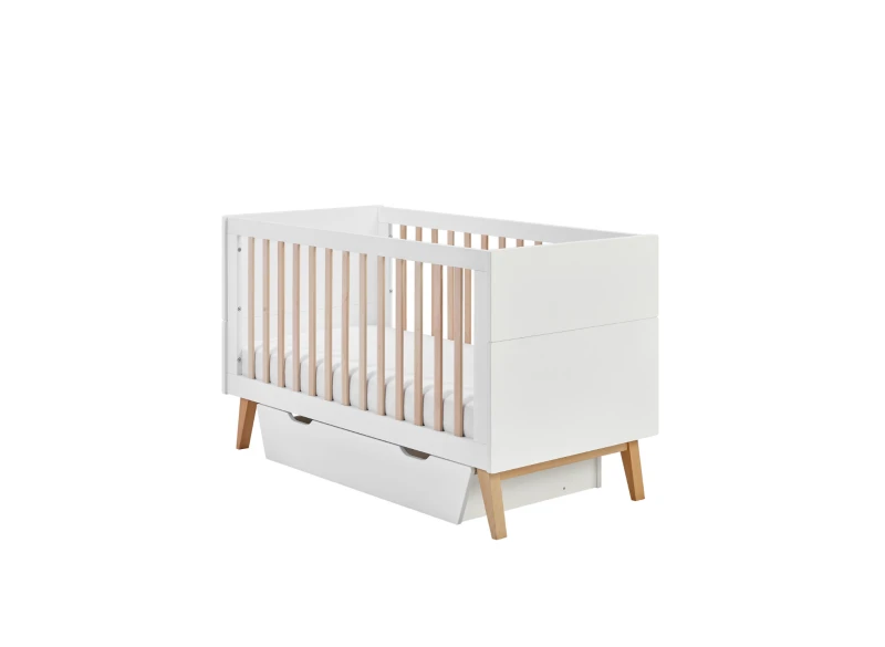 Białe łóżeczko niemowlęce ze szczebelkami i wysuwaną szufladą.