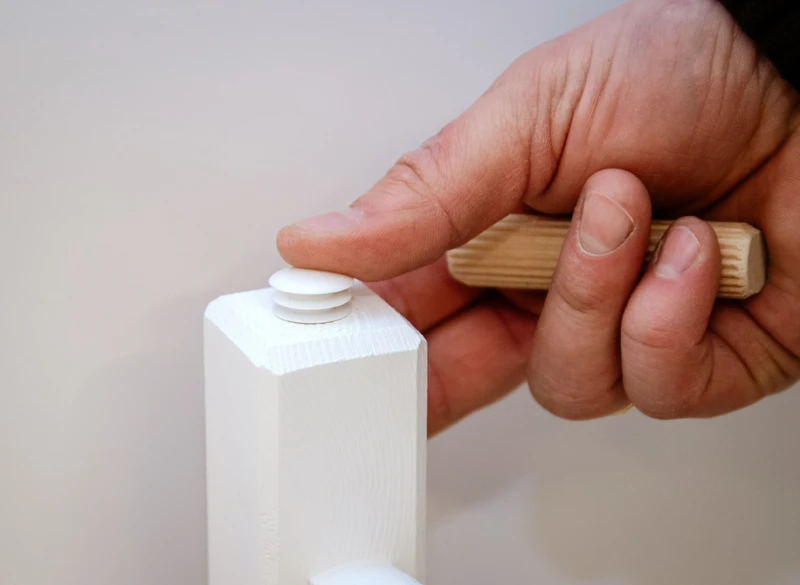 Ręczne wkładanie białej zaślepki w otwór montażowy ramy łóżka z litego drewna.