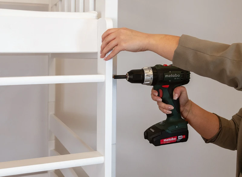 Montaż szczebla drabinki do białego łóżka piętrowego przy użyciu wkrętarki.