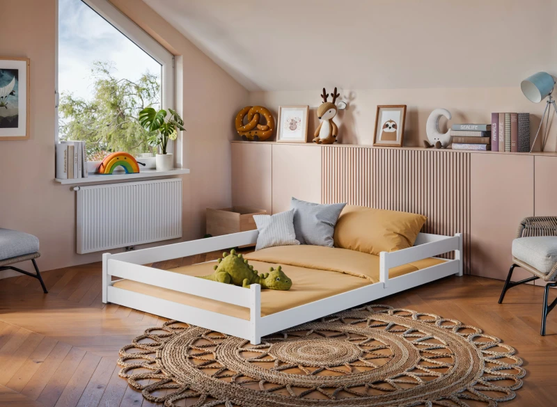 Białe drewniane łóżko podłogowe Montessori z żółtym materacem w przytulnym pokoju dziecięcym z dywanem.