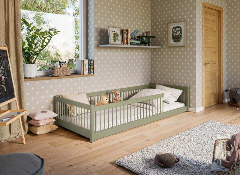 Niskie łóżko dziecięce z barierkami, oliwkowe drewno, z materacem i pluszakami, w pokoju z tapetą w kropki.