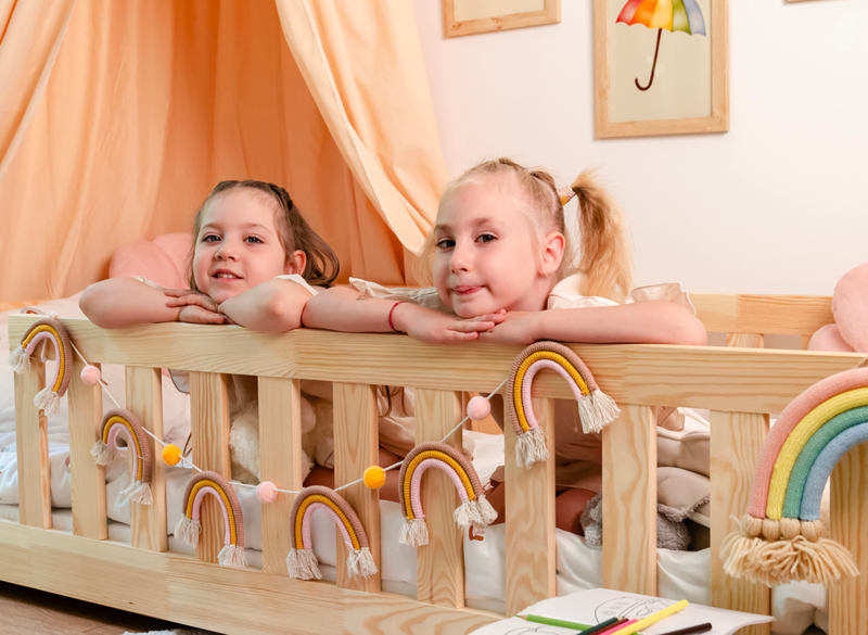 Dzieci wyglądające zza barierki drewnianego łóżeczka ozdobionego tęczowymi girlandami.