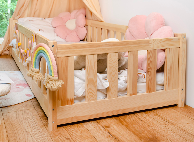 Drewniane łóżko dziecięce z barierką, różowymi poduszkami kwiatami i ozdobną tęczą.