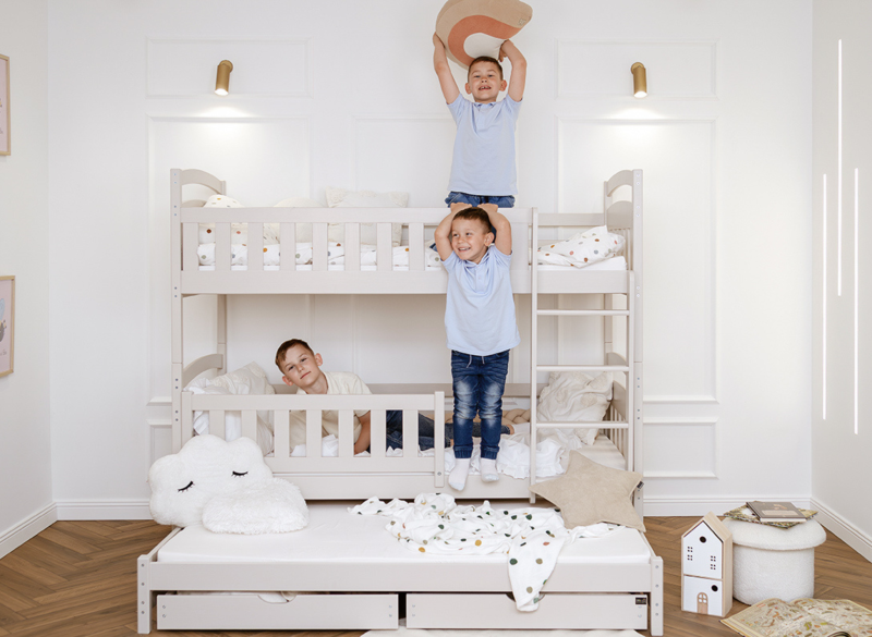 Jasnoszare drewniane łóżko piętrowe potrójne z dostawką i szufladami, z bawiącymi się chłopcami w nowoczesnym pokoju.