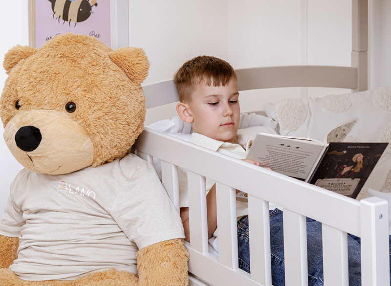 Chłopiec czytający książkę w białym łóżku piętrowym obok dużego misia.