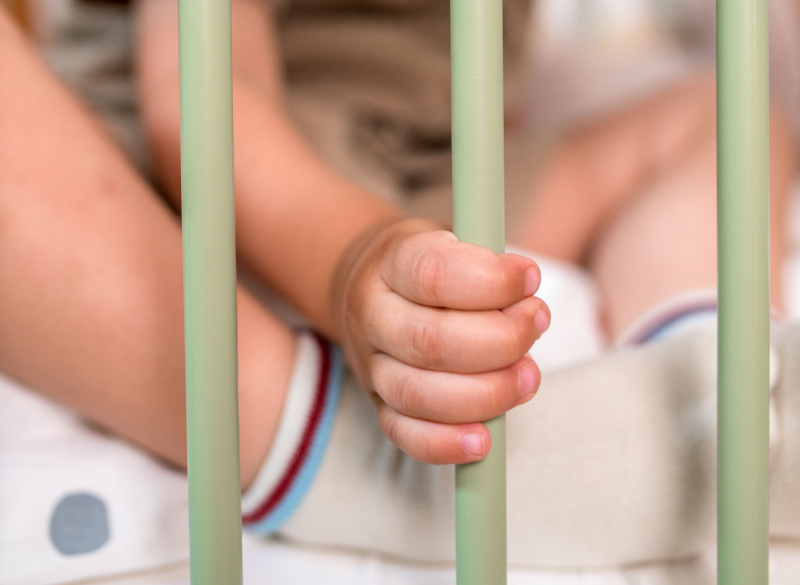 Mała rączka dziecka trzymająca się szczebelków zielonego łóżeczka niemowlęcego.