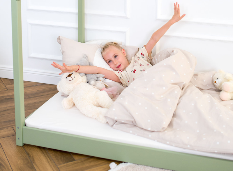 Dziewczynka rozciągająca się w zielonym drewnianym łóżku dziecięcym z beżową pościelą w kropki, w aranżacji pokoju.