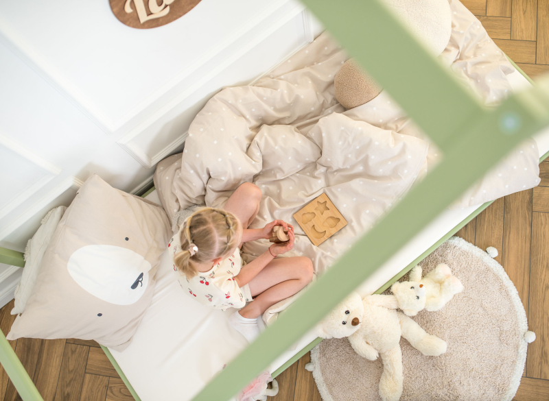 Dziewczynka bawi się drewnianą układanką na zielonym łóżku typu domek.