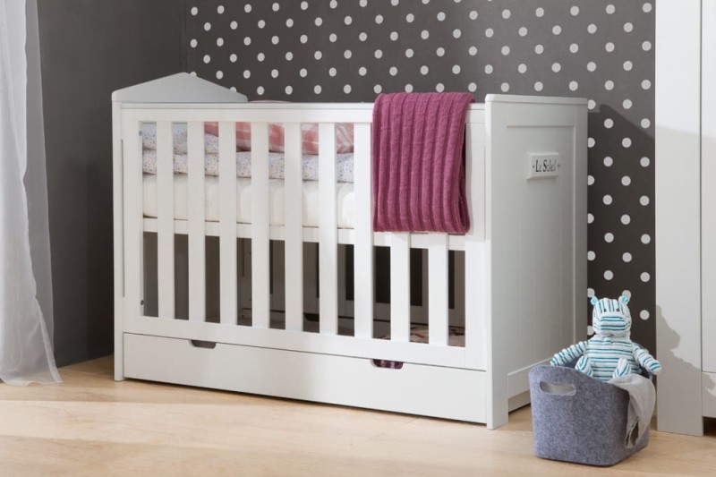 Białe łóżeczko niemowlęce z szufladą ustawione przy ścianie w kropki.