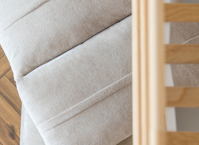 Materac beżowy z pikowaną tkaniną, widoczne przeszycia i drewniana rama łóżka, na tle drewnianej podłogi.