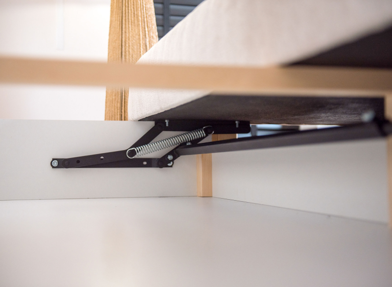 Mechanizm podnoszenia łóżka z czarnymi ramionami i sprężyną, widoczny w białym pojemniku na pościel.