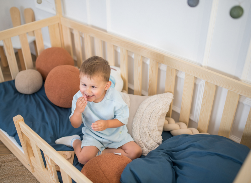 Mały chłopiec siedzi na łóżku domku z naturalnego drewna sosnowego z niebieskim materacem.