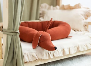 Rudy ochraniacz jamnik ułożony w łóżeczku z beżową pościelą.