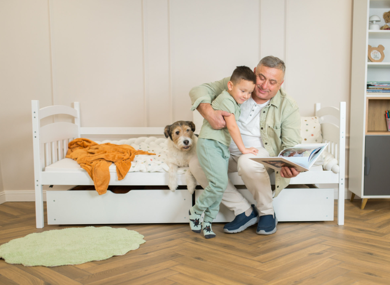 Dziadek z wnukiem i psem siedzący na białym łóżku i oglądający wspólnie książkę.
