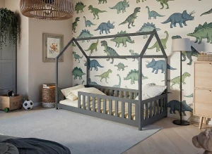 Szare łóżko domek dla dziecka na tle tapety z dinozaurami w przytulnym pokoju.