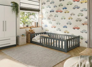 Szare łóżko parterowe w pokoju dziecięcym z tapetą w samochody i białą szafą.