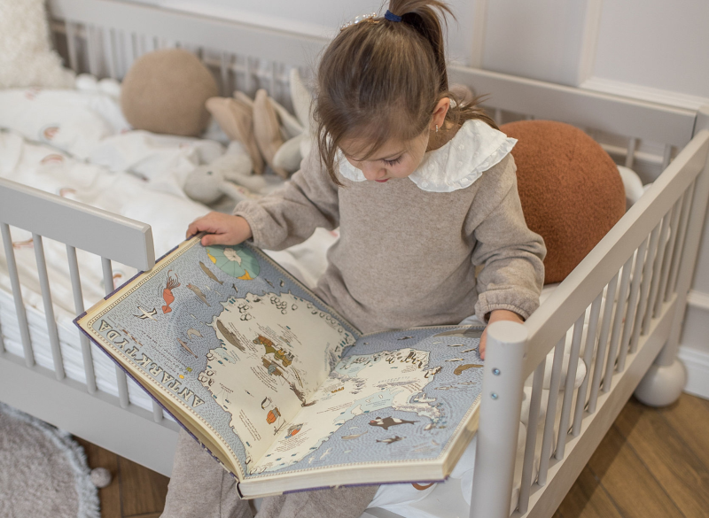 Mała dziewczynka siedzi na łóżku i czyta dużą książkę edukacyjną o Antarktydzie.