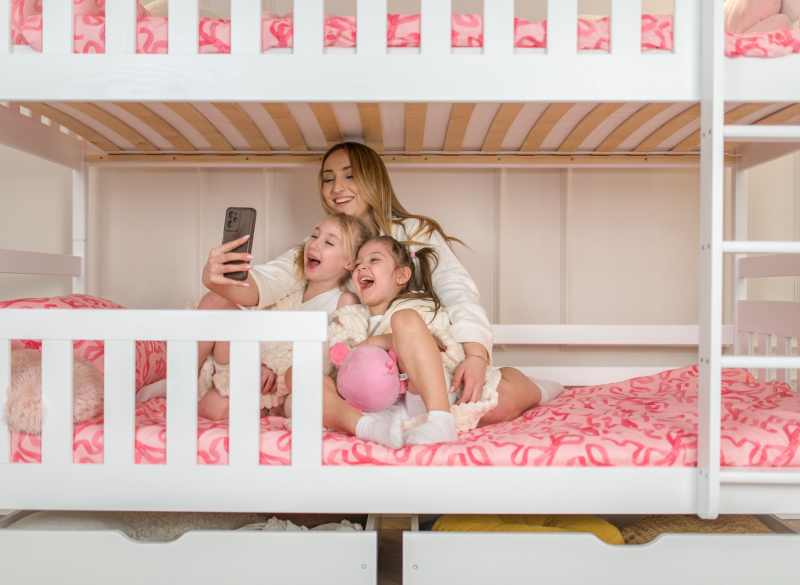 Wesołe selfie mamy i córek na dolnym poziomie białego łóżka piętrowego.