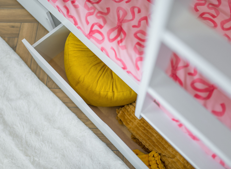 Wysunięta biała szuflada pod łóżko z żółtą pufą i kocykiem w pokoju dziecka.