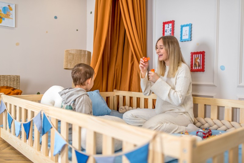 Mama puszcza bańki mydlane synkowi siedzącemu na drewnianym łóżku z niebieską pościelą.