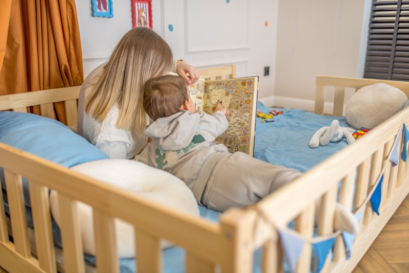 Mama i syn czytają dużą ilustrowaną książkę na niskim łóżku z litego drewna.