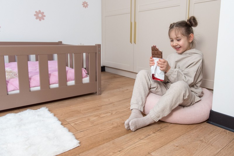 Uśmiechnięta dziewczynka siedzi na różowej pufie na podłodze, trzymając nadgryzioną tabliczkę czekolady. Obok widoczne niskie łóżko w kolorze mlecznej czekolady z różową pościelą. W tle beżowa szafa.