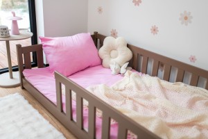 Niskie, drewniane łóżko dziecięce z barierką ochronną w kolorze mlecznej czekolady. Na łóżku leży różowa pościel i jasny, ażurowy kocyk, a obok barierki stoi poduszka w kształcie białego kwiatu. W tle widoczna jest biała ściana w różowe wzory kwiatowe oraz fragment beżowego stolika.